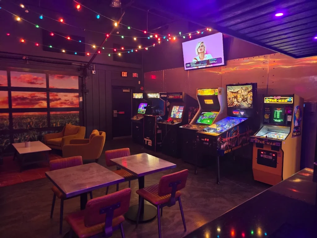 Interior of The Life Bar Arcade area with tables for patrons to sit down surrounde dby arcade games and pinball machines in colorful lighting.